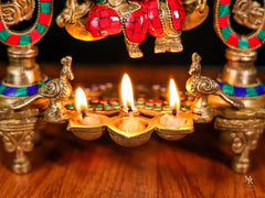 Three diya lamps on tray with small flames, decorative base with Radha Krishna swing details