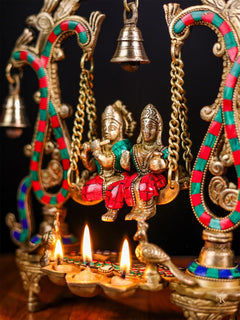 Close-up of Radha Krishna swing idol, gold chains, hanging bell, diya flames below