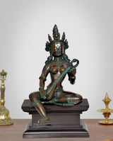 Brass Saraswati idol seated on pedestal, holding veena; dark green patina finish