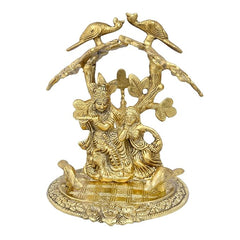 Gold-tone Radha Krishna idol under tree with birds, front view on textured base