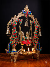 Radha Krishna statue on swing with two seated figures, hanging bell, diya flames