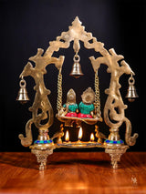 Radha Krishna swing statue with hanging bell and diya flames, gold arch and chains