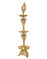 Gold-toned brass diya stand with peacock finial above three stacked oil-lamp plates