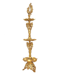 Gold-toned brass diya stand with peacock finial above three stacked oil-lamp plates