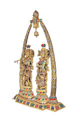 Radha and Krishna figurines under brass arch with colorful accents, bell