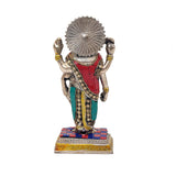Dhanvantari brass statue from behind; crown and fan-like halo, red robe with teal accents.