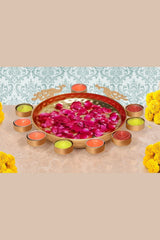 Red peacock urli bowl with floating pink petals, orange tea lights, and yellow blooms