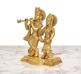 Brass Radha Krishna idol—Krishna raises flute, Radha holds bowl; peacock beside.
