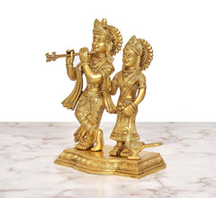 Brass Radha Krishna idol—Krishna raises flute, Radha holds bowl; peacock beside.
