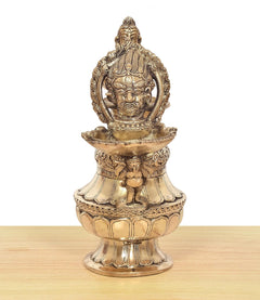 Brass oil lamp with standing deity figure holding a lamp bowl, ornate relief base