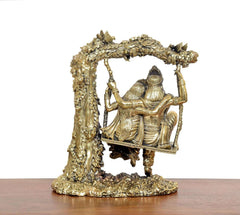 Brass Radha-Krishna idol on swing, rear view with frame arch and hanging chains.