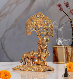 Metal Krishna figurine playing flute beside cow idol, under filigree tree canopy