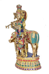 Brass Krishna figurine beside cow, richly painted robe, jewelry, and decorative halo