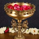 Brass urli stand with wide bowl and detailed deity base; bowl holds red petals
