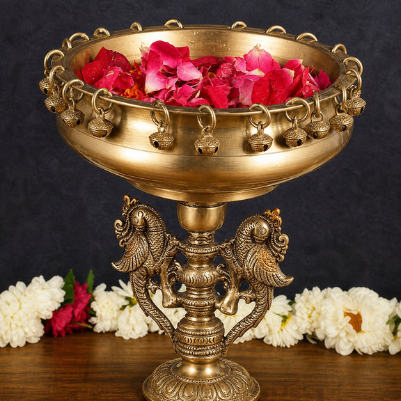 Brass urli stand with wide bowl and detailed deity base; bowl holds red petals
