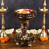 Brass urli centerpiece on ornate pedestal with chain rim holding flowers and side diyas