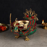 Decorative brass urli with peacock top, teal and red enamel bands, lit candle in center