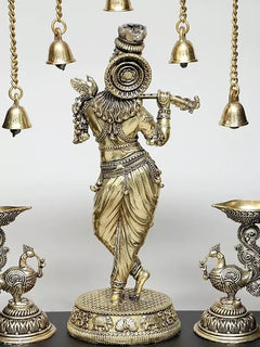 Brass Lord Krishna in profile with raised hand, hanging bells, and lamp bowls in frame