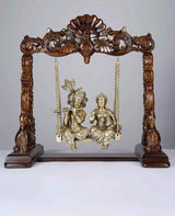 Brass Radha-Krishna statue on wooden swing with ornate posts and hanging chains