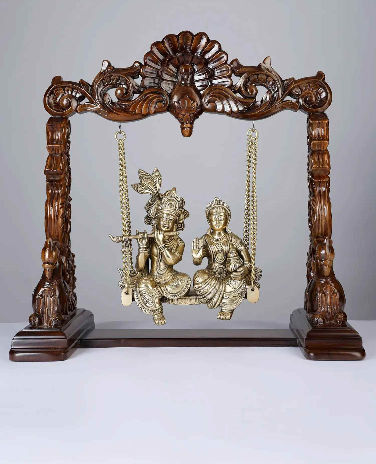Brass Radha-Krishna statue on wooden swing with ornate posts and hanging chains