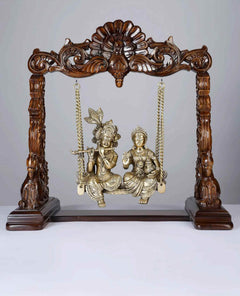 Brass Radha-Krishna statue on wooden swing with ornate posts and hanging chains