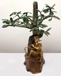 Brass Krishna playing flute figure beside twisting dark tree trunk and leaf canopy