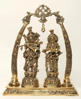 Brass Radha Krishna statue on tusk-style stand, ornate hanging bells and arch