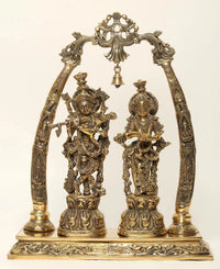 Brass Radha Krishna statue on tusk-style stand, ornate hanging bells and arch