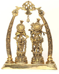 Brass Radha Krishna statue with carved arched frame and two figurines, bell hanging