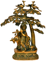 Brass Krishna playing flute statue under mango tree, decorative leaves and base